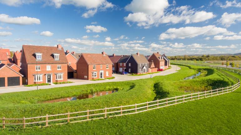 Row of modern brick houses along a curved road with green fields and a wooden fence.